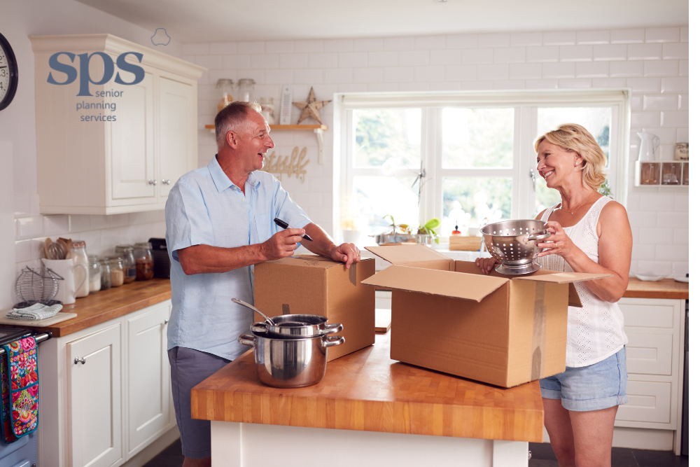 Senior man and his daughter smiling while packing boxes in a kitchen, preparing to downsize from their family home with the help of senior planning services.